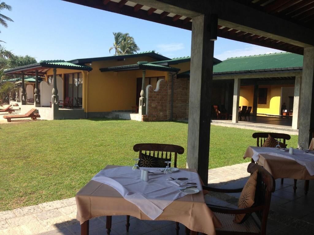 A covered outdoor area with tables and chairs, a grassy lawn, and buildings with yellow walls and green roofs.