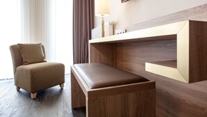 Una habitación de hotel moderna con suelo de madera, un sillón marrón, un banco de madera, un televisor montado en la pared y una mesita de noche con lámpara.