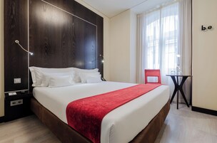 Hotellihuone, jossa on suuri sänky, punainen heittopeitto, pieni pöytä ja tuoli.