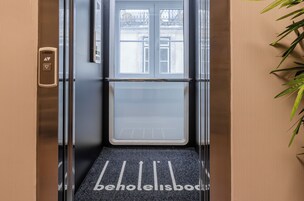An elevator with a glass door, a metal handrail, and a black mat with "behotelisboa" text.