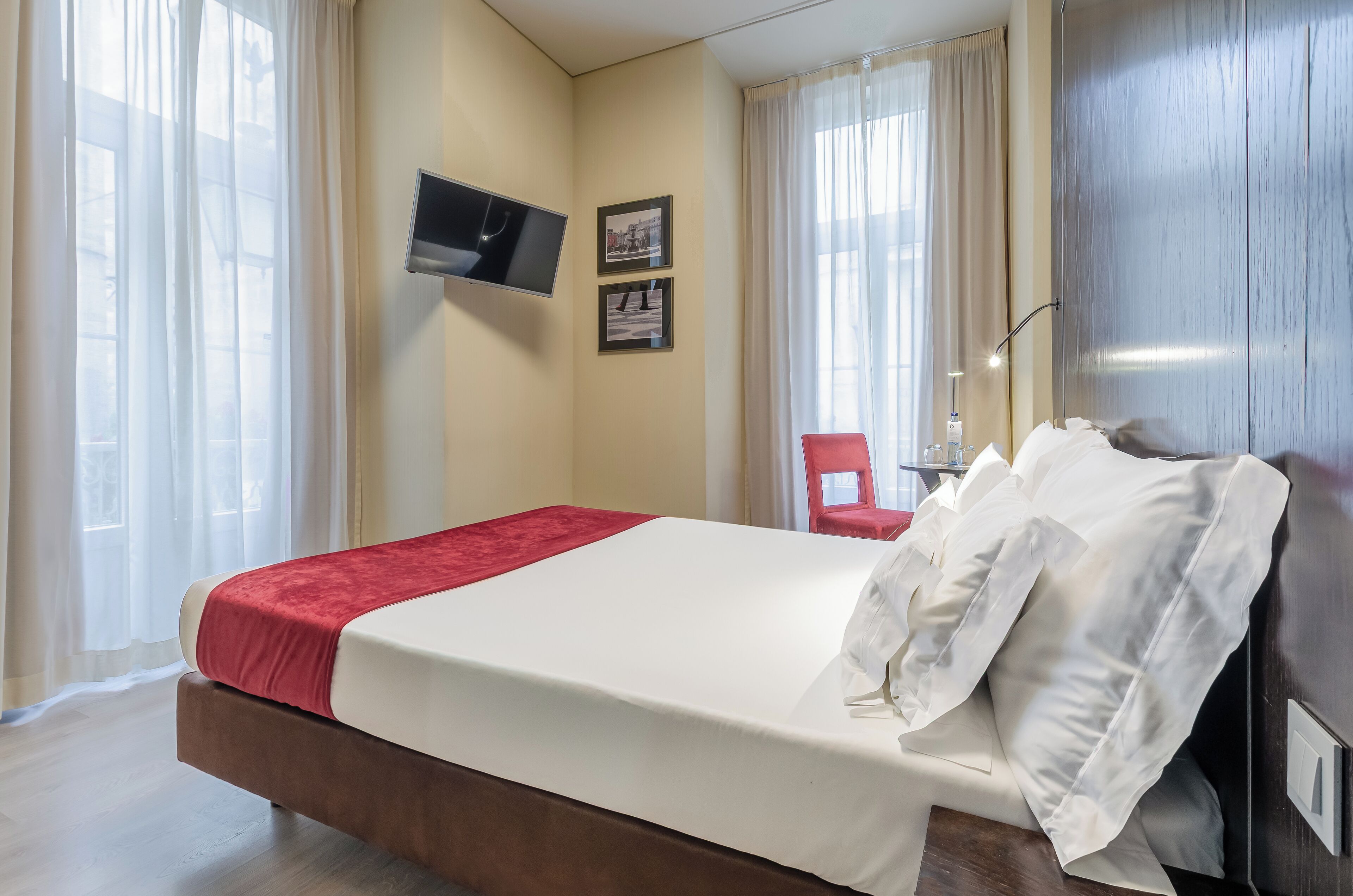 A hotel room with a bed, a TV mounted on the wall, and a window with curtains.