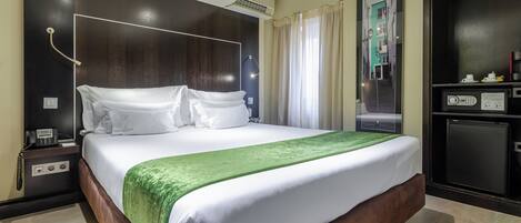 A hotel room with a large bed, a bedside table with a phone, a mirror, and a small refrigerator.