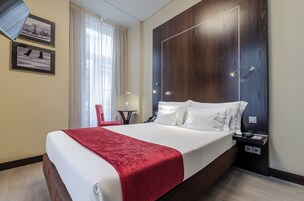 A hotel room with a large bed, a red throw blanket, a wooden headboard, a nightstand with a phone, and a window with curtains.