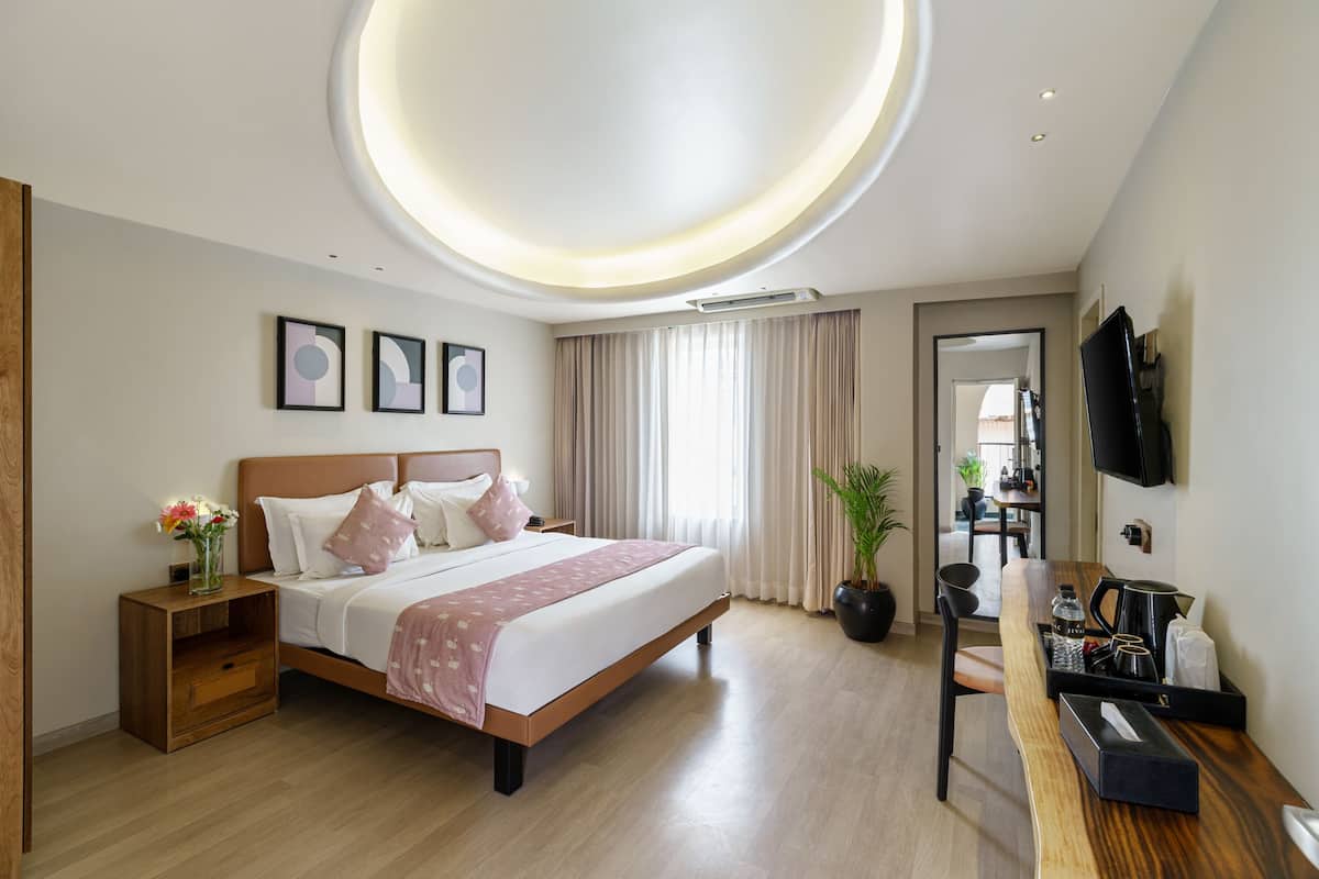 A modern hotel room with a large bed, bedside tables, a desk with a chair, a TV mounted on the wall, and a circular ceiling light.