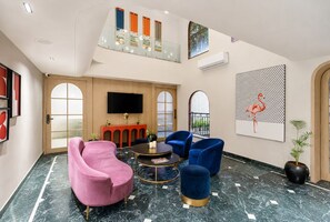 A modern lobby with a marble floor, a pink armchair, blue armchairs, a round table, and a TV mounted on the wall.