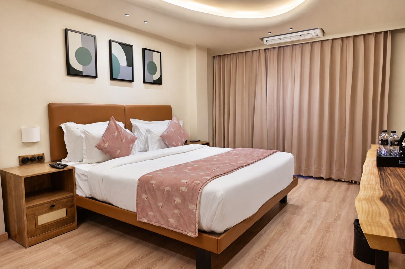 A hotel room with a bed, bedside table, lamp, artwork, curtains, and a wooden table with bottles.