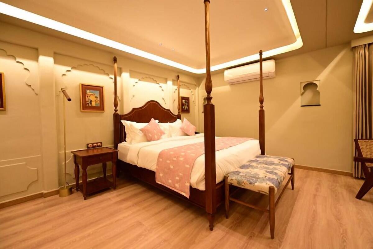 A four-poster bed with a wooden headboard, a bedside table, a bench, and a chair in a hotel room.