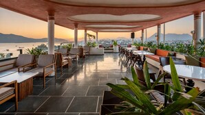 An outdoor seating area with tables and chairs, potted plants, and a view of the city and water. - My Downtown - The Red Pier (Udaipur)