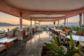 An outdoor seating area with tables and chairs, potted plants, and a view of the city and water. - My Downtown - The Red Pier (Udaipur)