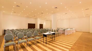 Un salón de conferencias con filas de sillas y mesas, suelo de madera y luces empotradas en el techo.