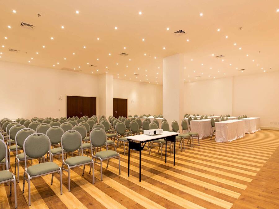 Un salón de conferencias con filas de sillas y mesas, suelo de madera y luces empotradas en el techo.