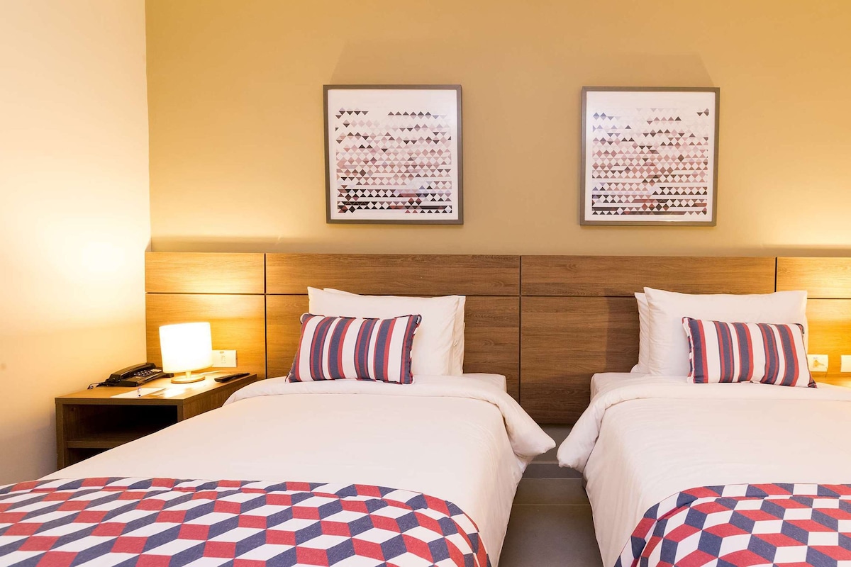 Une chambre d’hôtel avec deux lits, une tête de lit en bois, des lampes de chevet et une œuvre d’art abstraite encadrée accrochée au mur.