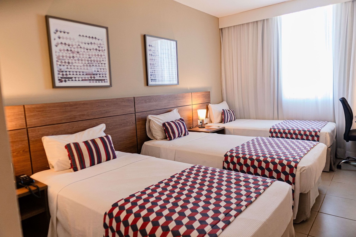 Une chambre d’hôtel avec deux lits, chacun recouvert d’une couvre-lit à motifs, une table de chevet avec une lampe et une grande fenêtre avec des rideaux.