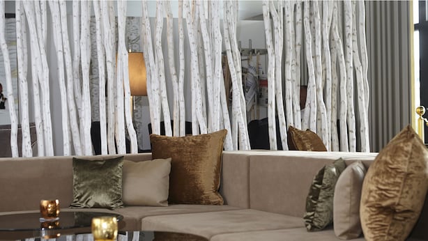 A modern living room with a beige sofa, multiple cushions, a glass coffee table, and a decorative white tree-like structure. - Hotel AVA (Neuenkirch)