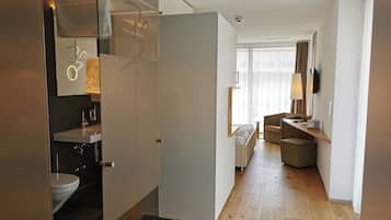 Habitación de hotel moderna con suelo de madera, baño con inodoro y lavamanos, cama, una pequeña zona de estar y un ventanal grande con cortinas.