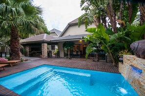 Piscina con caída de agua, rodeada de plantas tropicales y un patio con mobiliario exterior.