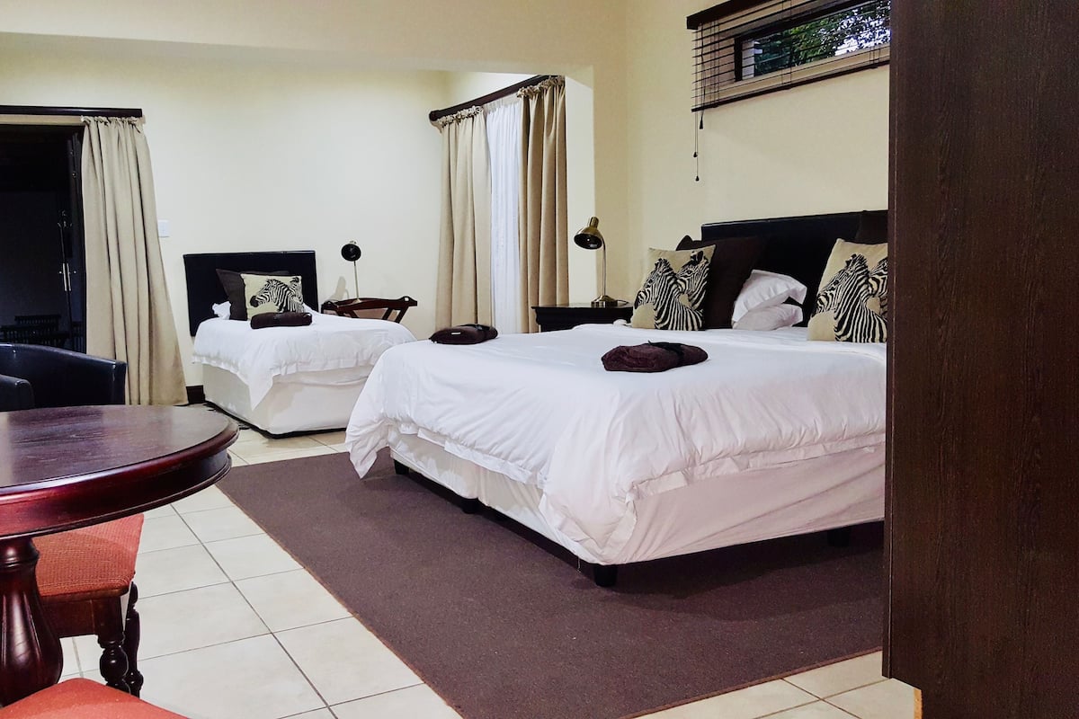 A hotel room with two beds, a small table with a chair, a potted plant, and a window with curtains.