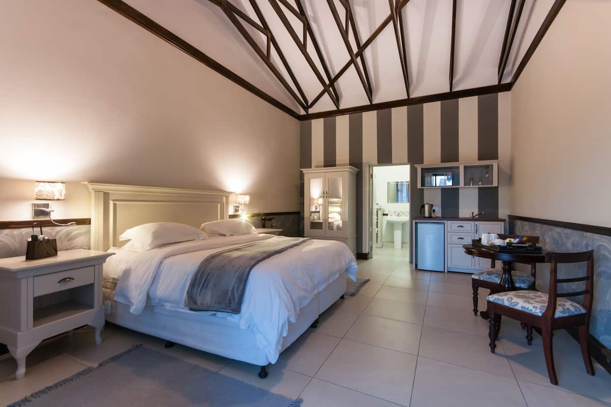 A bedroom with a bed, bedside tables, a desk, and a chair. There is a kitchenette with cabinets and a refrigerator.