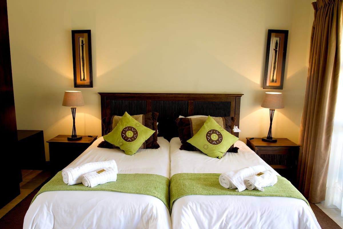 A hotel room with two beds, each with a green bedspread, white linens, and matching pillows. There are two bedside tables with lamps, a wooden headboard, and framed artwork on the walls.