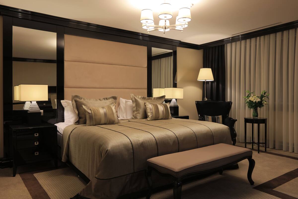 A hotel room with a large bed, a bench, a nightstand with a lamp, a mirror, and a small table with a vase.