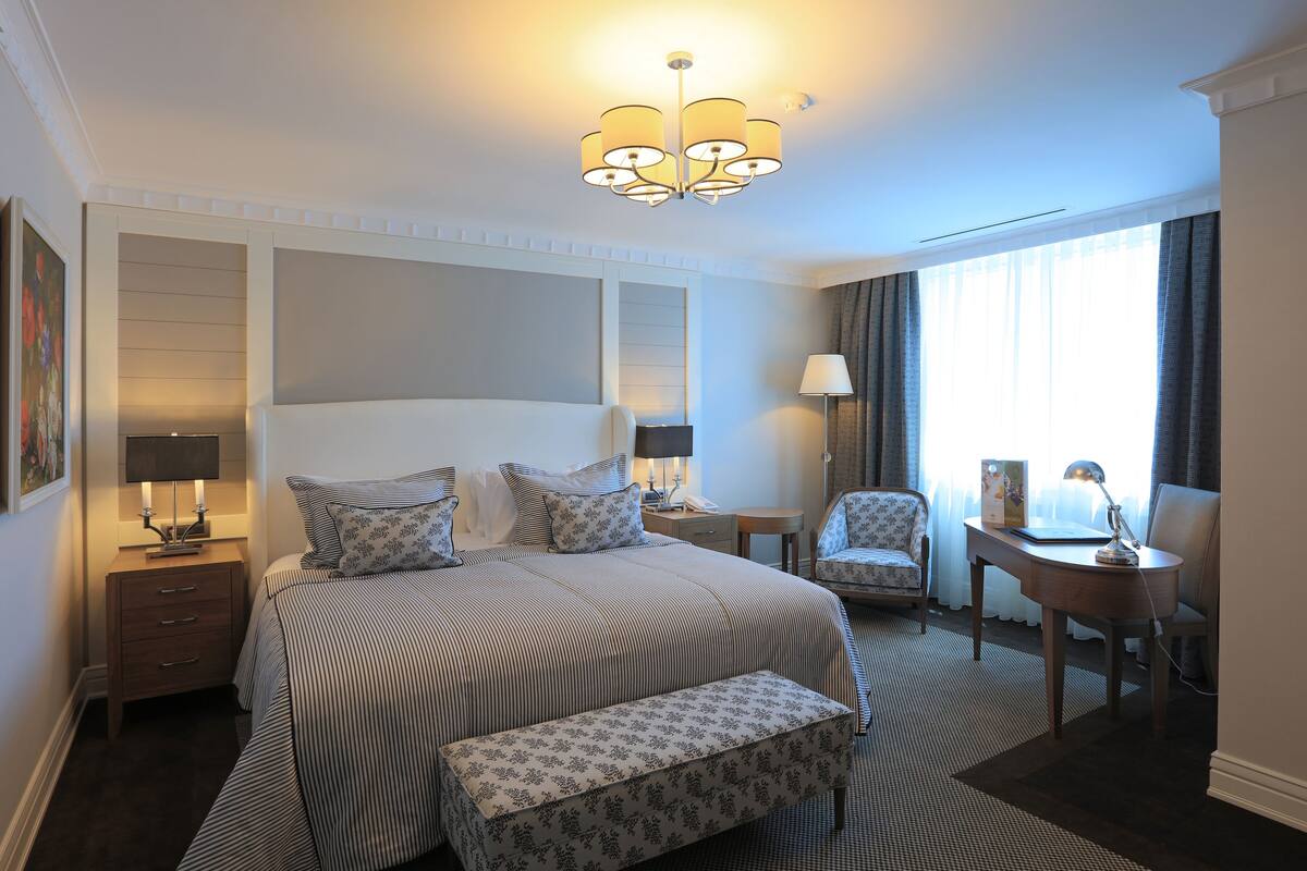 A hotel room with a large bed, bedside tables, a chair, a desk, and a chandelier.