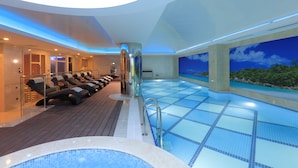 A modern indoor swimming pool with a wooden deck and lounge chairs, a hot tub, and a large mural of a tropical landscape.