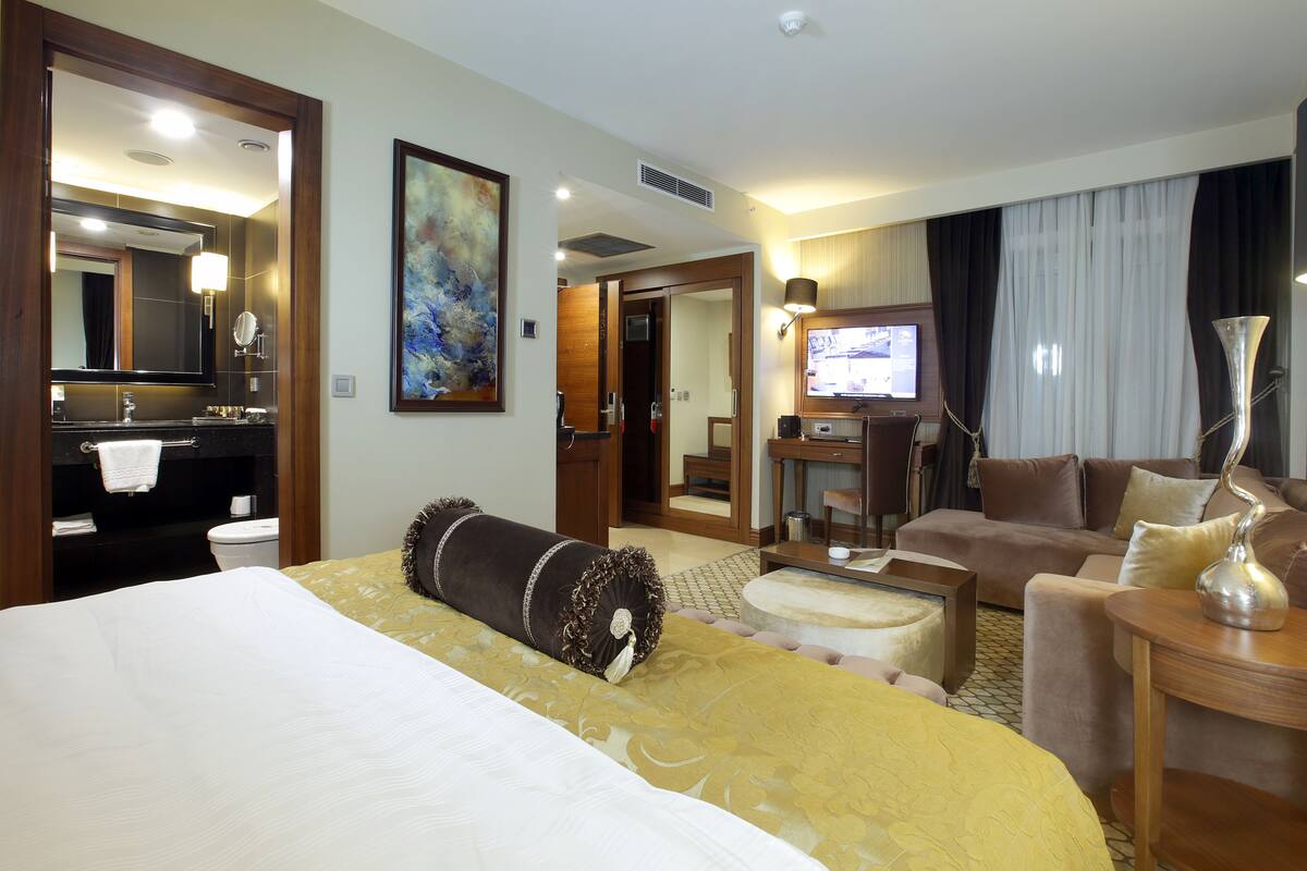 A hotel room with a large bed, a desk with a computer, a sofa, a TV, and a bathroom with a mirror and sink.