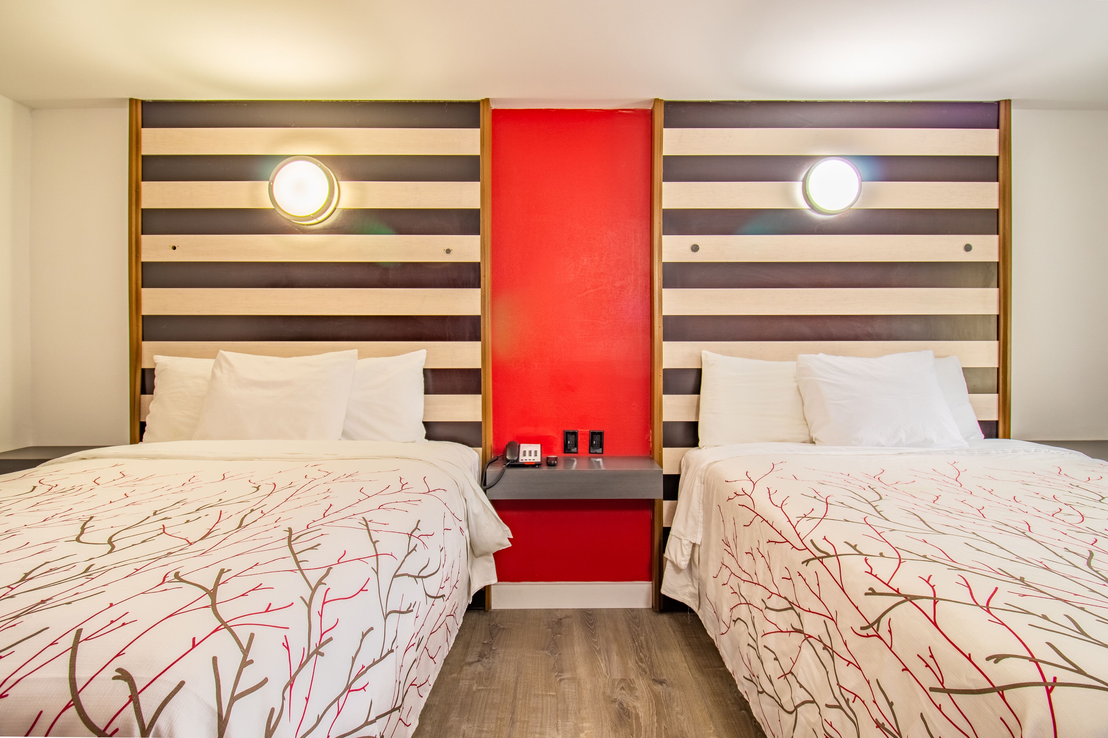 Quarto de hotel com duas camas, uma parede de destaque vermelha e uma pequena mesa com telefone e luminária.