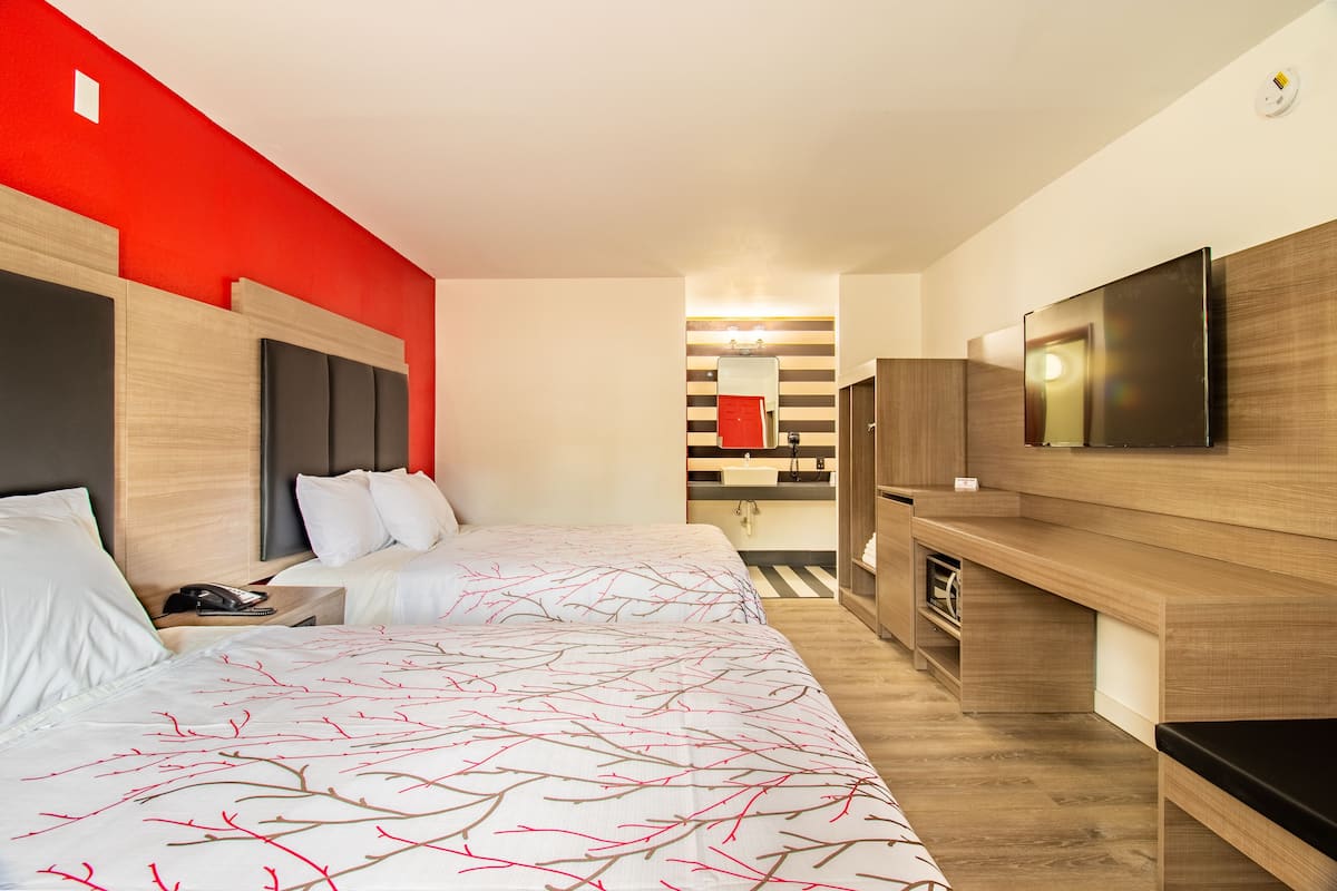 A hotel room with two beds, a TV, a desk, and a bathroom visible through a glass partition.