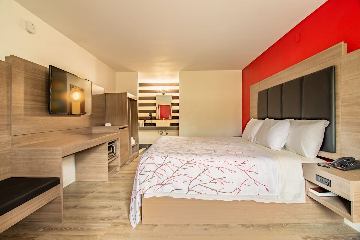 A modern hotel room with a wooden bed, a desk, a mirror, and a red accent wall.