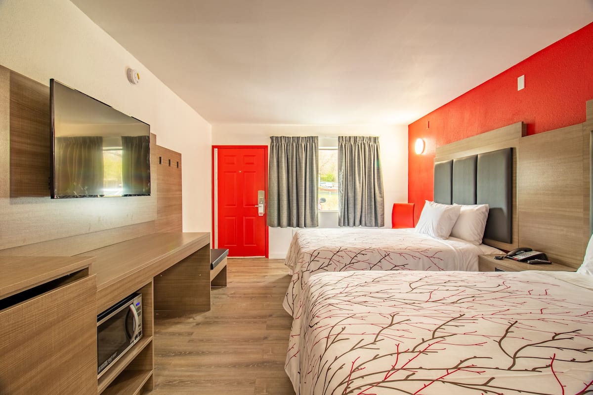 A hotel room with a flat-screen TV, two beds with patterned bedding, a red door, and a window with curtains.
