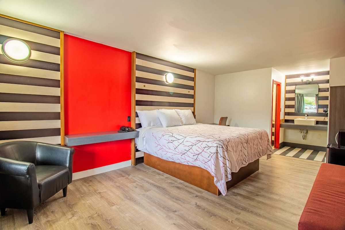 A modern hotel room with a large bed, a desk, a chair, and a bathroom with a mirror and sink.