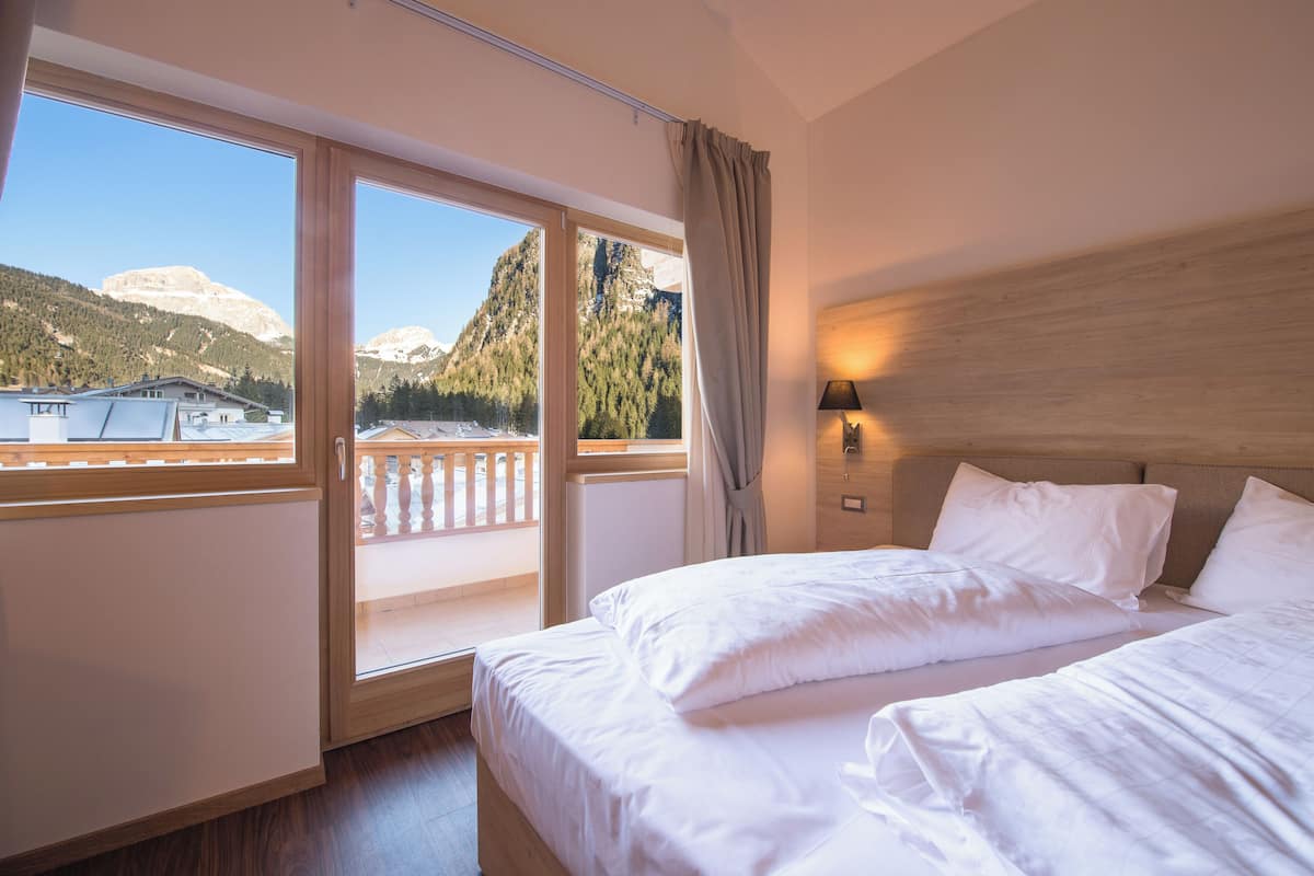 A hotel room with a large bed, a nightstand, a lamp, and a view of mountains through a large window.