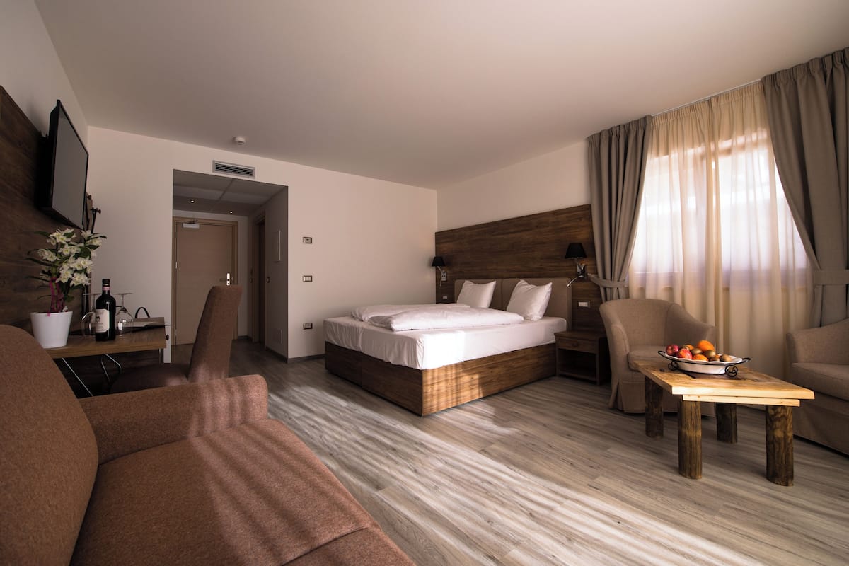 A hotel room with a bed, a sofa, a dining table with fruit, and a TV on the wall.