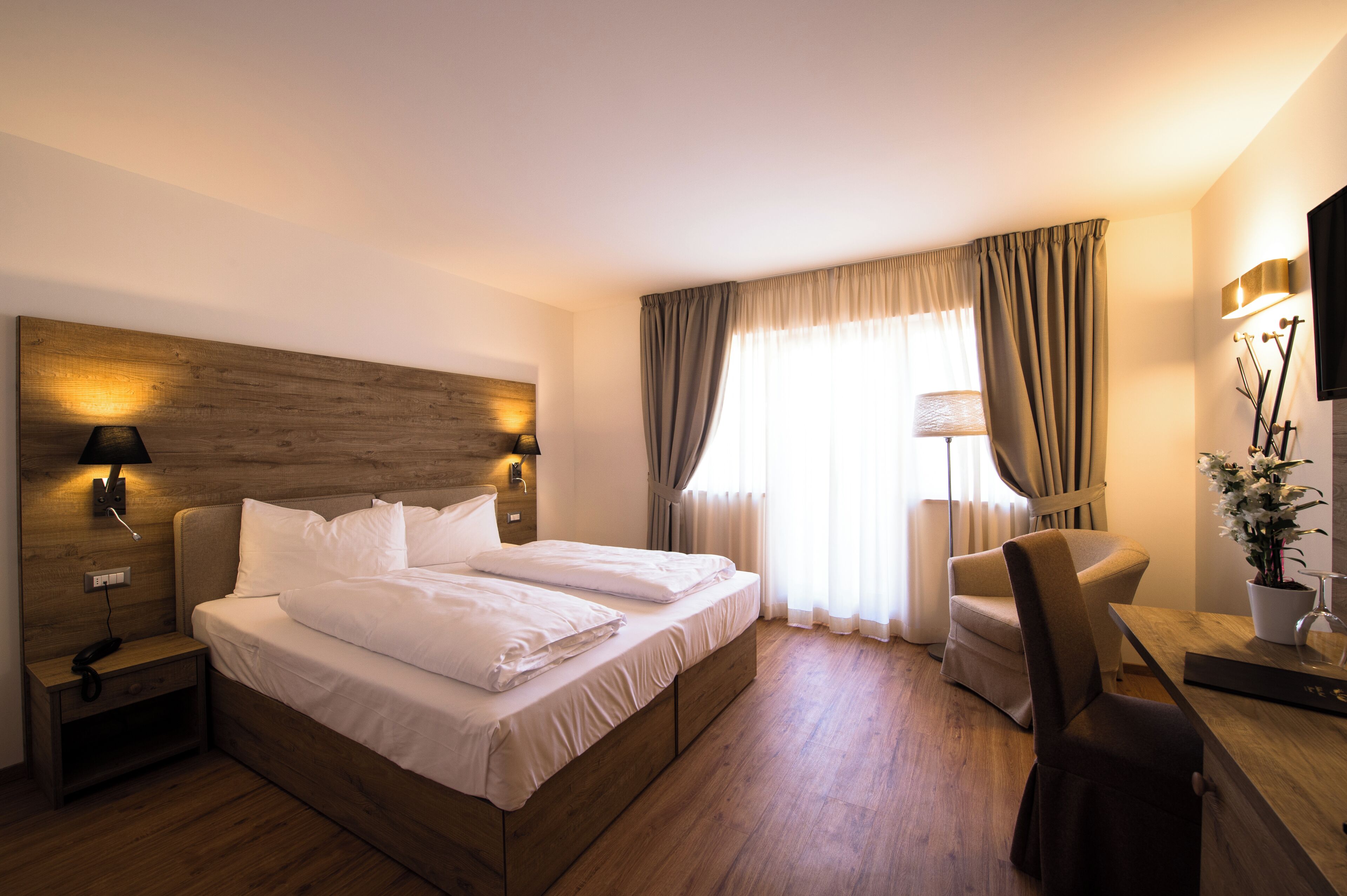 A hotel room with a wooden headboard, a bed with white linens, a bedside table with a lamp, a chair, and a TV mounted on the wall.