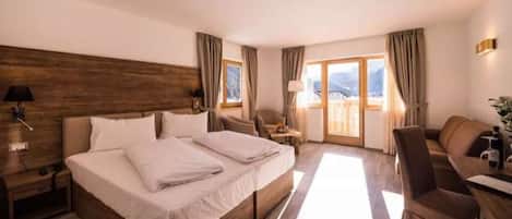 A hotel room with a large bed, a desk, a chair, and a view of mountains through the windows.