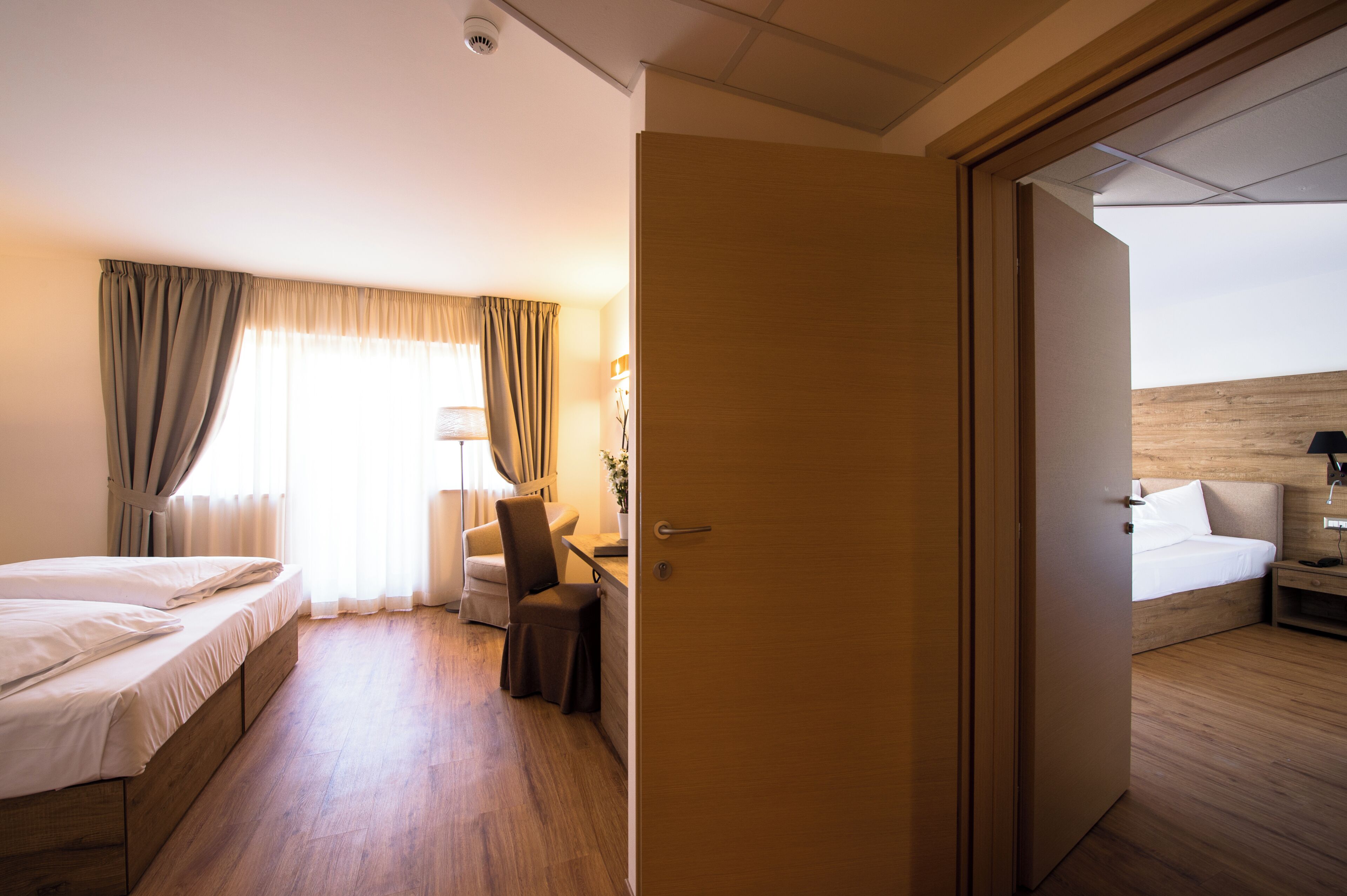 Quarto de hotel com cama, escrivaninha, cadeira e porta que dá para outro cômodo.