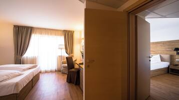 Quarto de hotel com cama, escrivaninha, cadeira e porta que dá para outro cômodo.