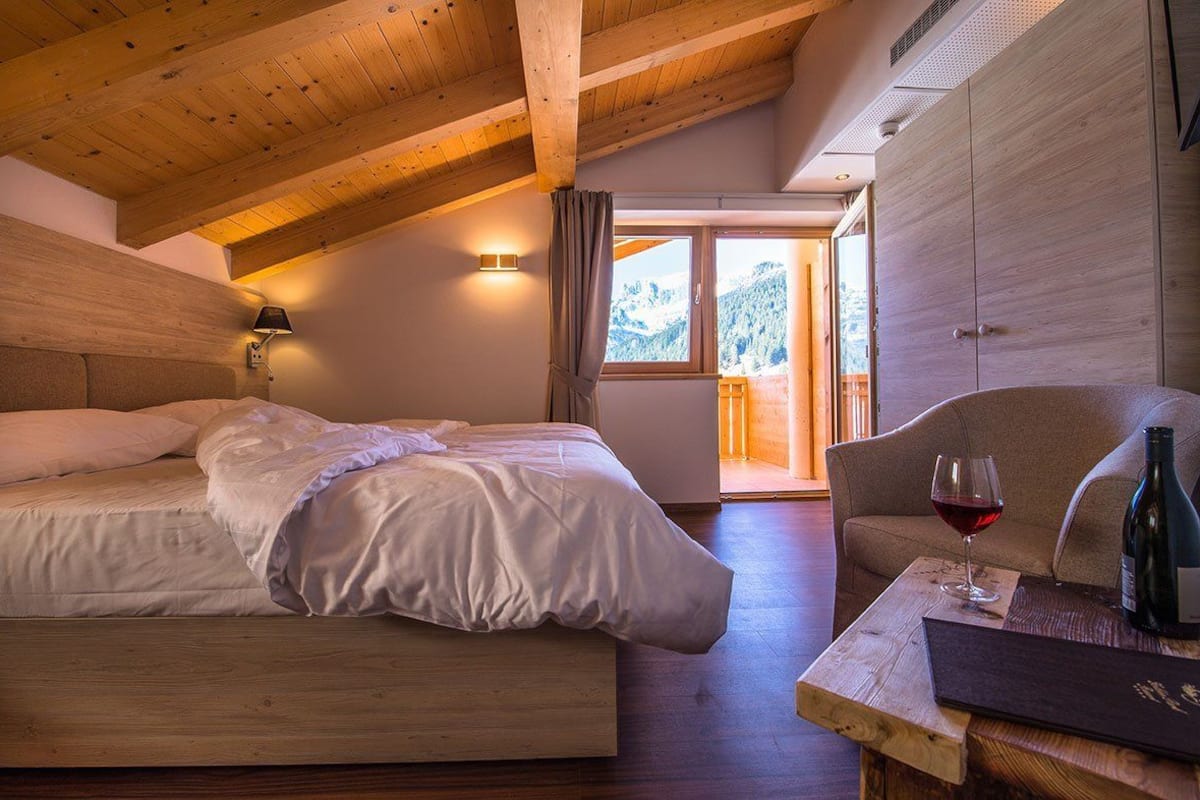 A bedroom with a wooden ceiling, a bed with white linens, a nightstand with a wine glass and bottle, and a view of a mountain through a window.