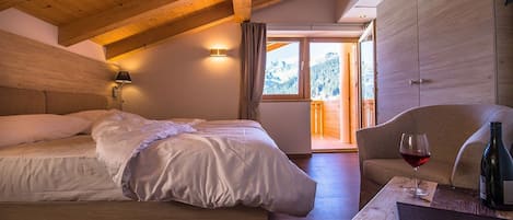 A bedroom with a wooden ceiling, a bed with white linens, a nightstand with a wine glass and bottle, and a view of a mountain through a window.