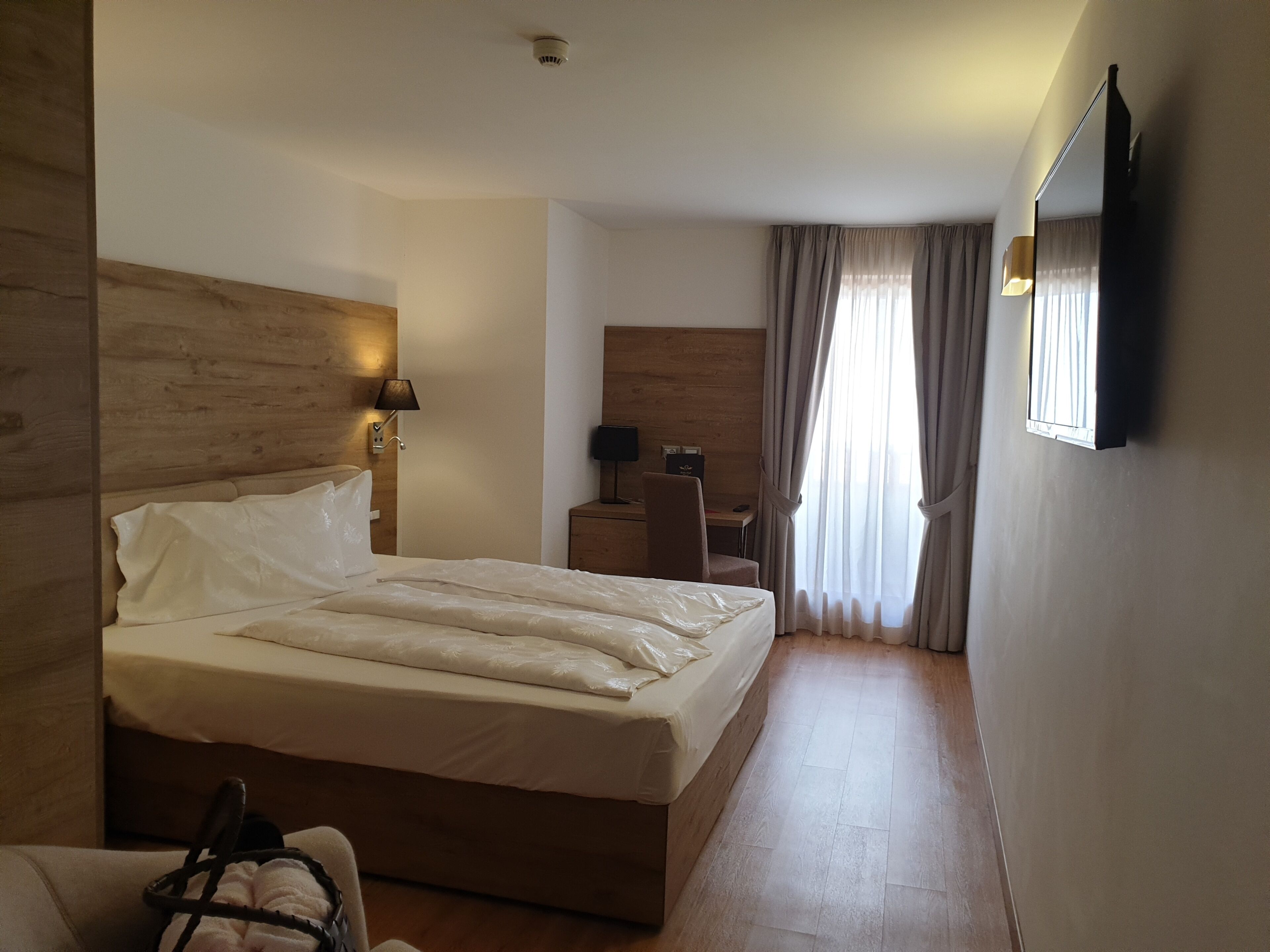 Une chambre d’hôtel avec un lit, un bureau, une chaise, une lampe, un miroir et une fenêtre avec des rideaux.