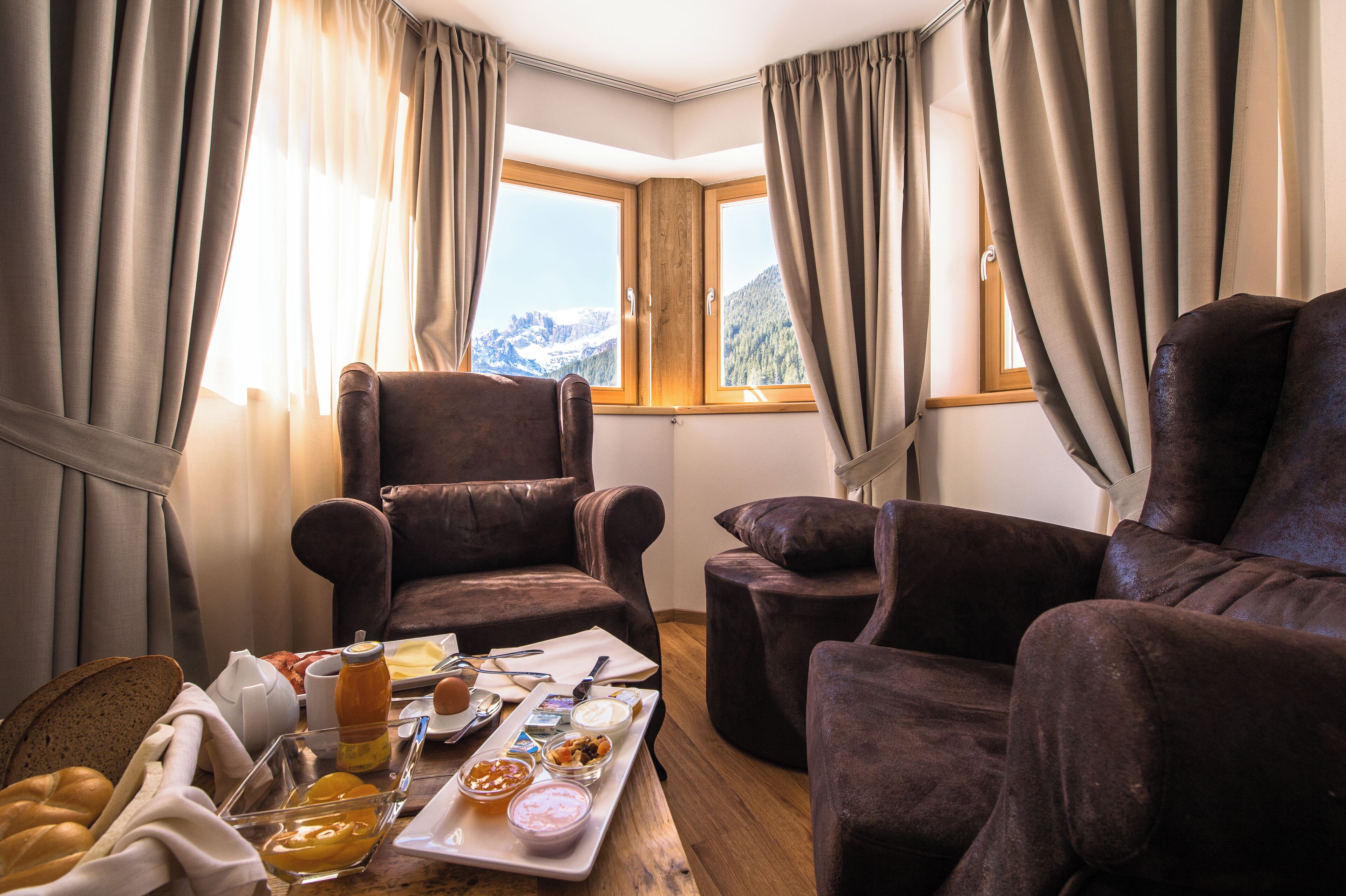 A room with a large window, two brown armchairs, a wooden table with breakfast items, and curtains.