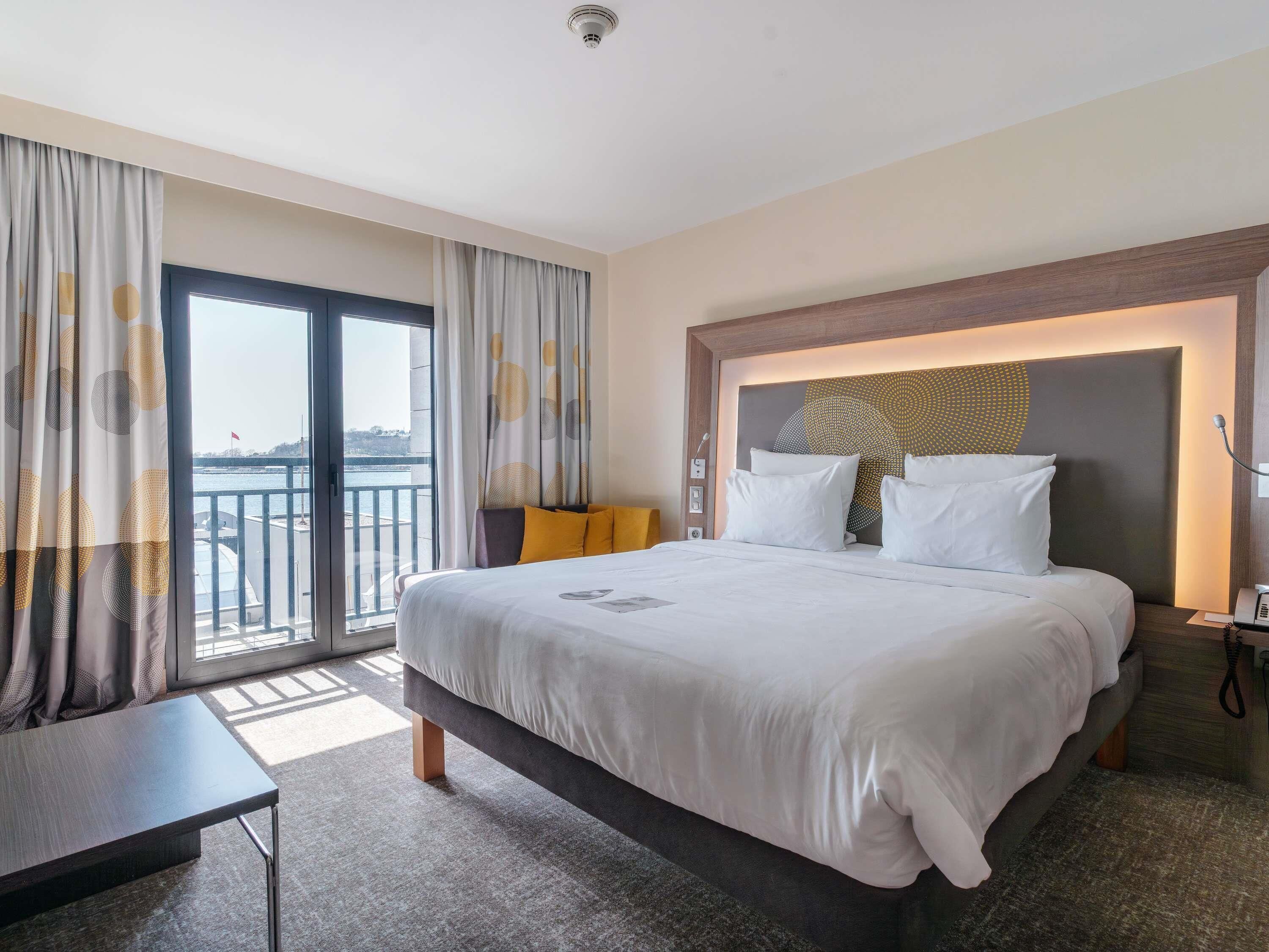 A hotel room with a large bed, a desk, a chair, a nightstand, and a view of the sea through the window.