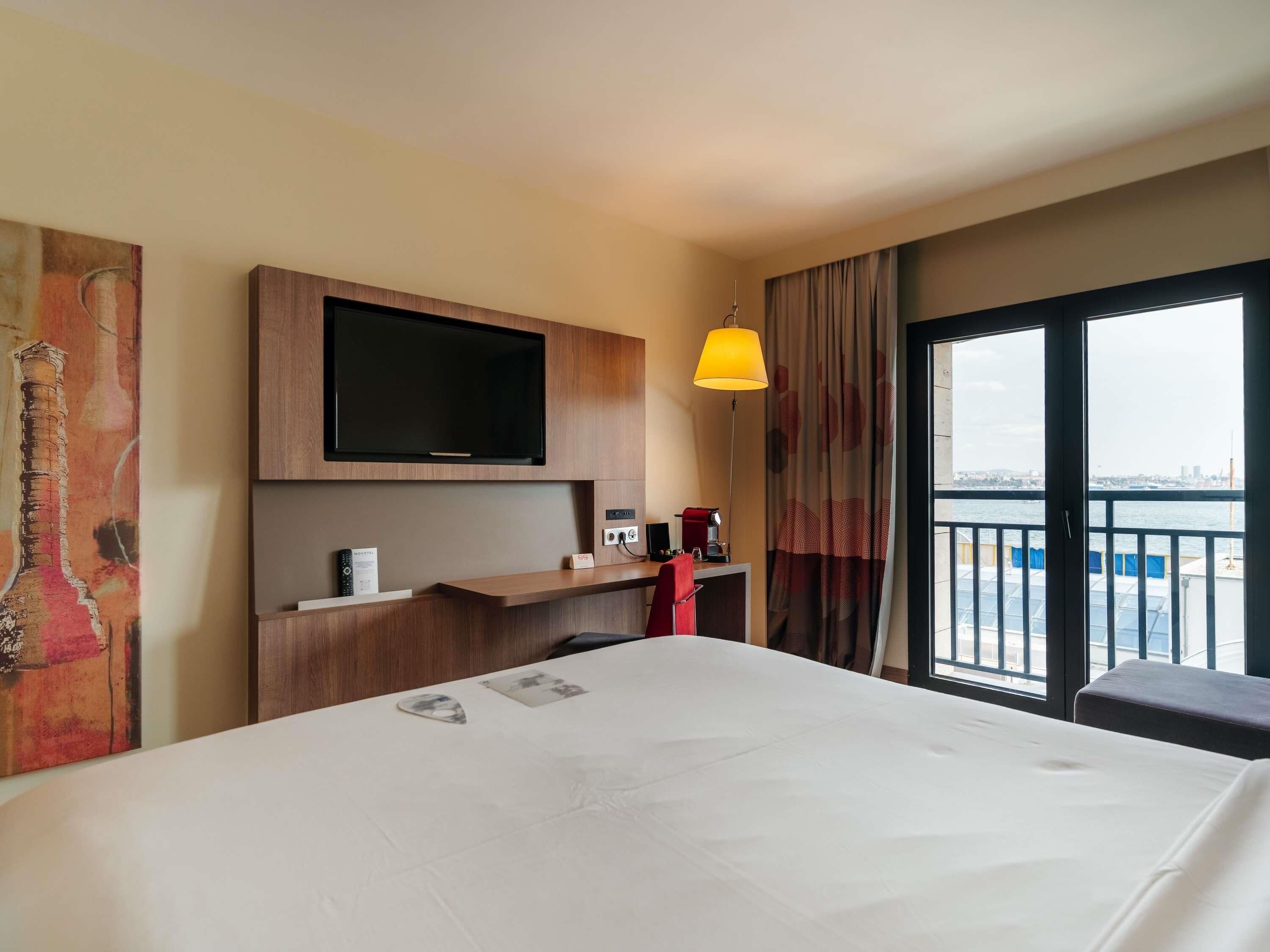 Habitación de hotel con cama, televisión montada en la pared, escritorio con lámpara y vista a la ciudad a través de un ventanal grande.