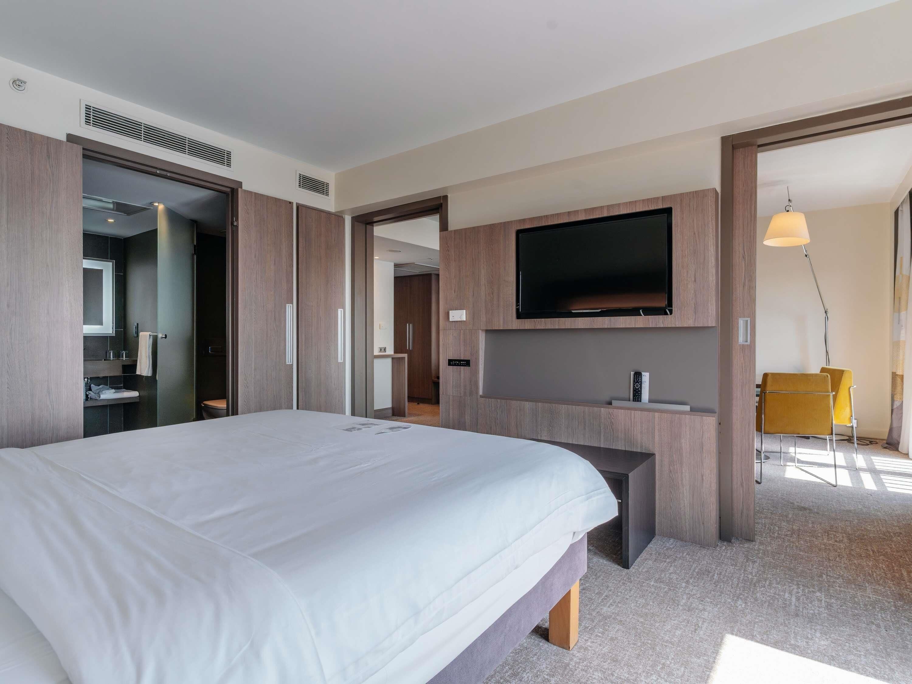 Una habitación de hotel moderna con una cama grande, un televisor de pantalla plana en la pared, un escritorio con silla, y un baño con espejo y lavamanos.