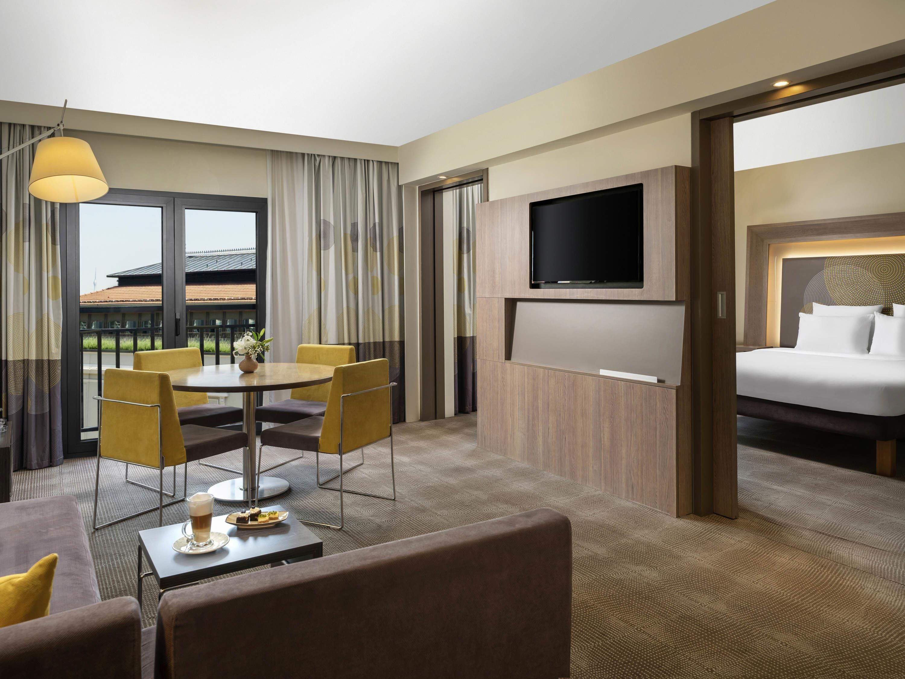 A hotel room with a bed, a TV, a dining area with chairs and a table, and a view through the window.