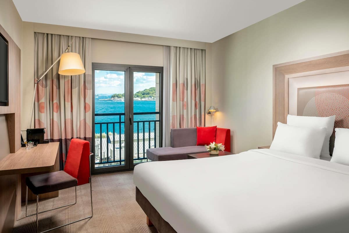 Une chambre d’hôtel comprenant un lit, un bureau, une chaise, un canapé, une télévision, une lampe, une fenêtre avec des rideaux et une vue sur la mer.