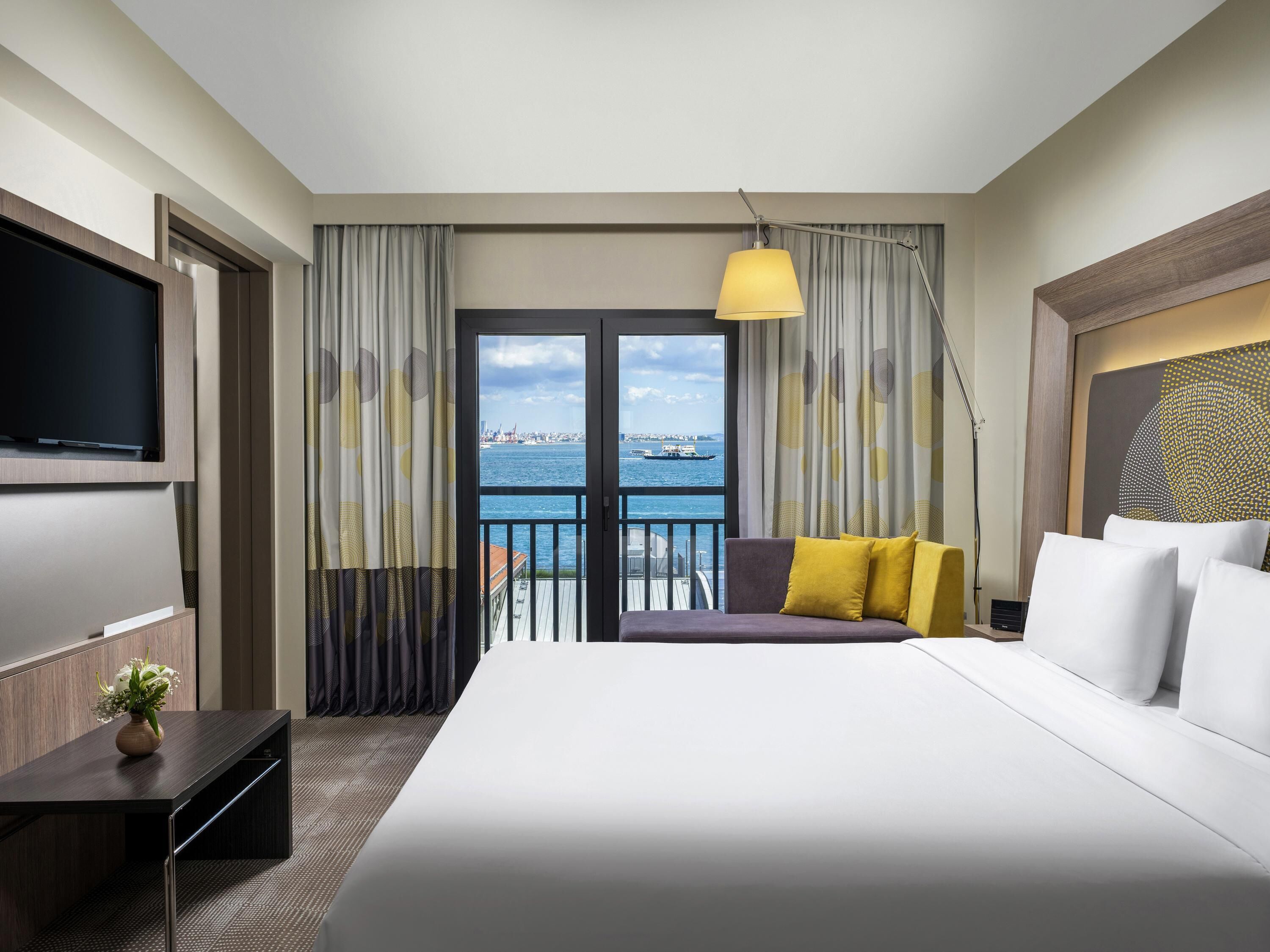 A hotel room with a large bed, a TV, a desk with a vase, and a view of the sea.
