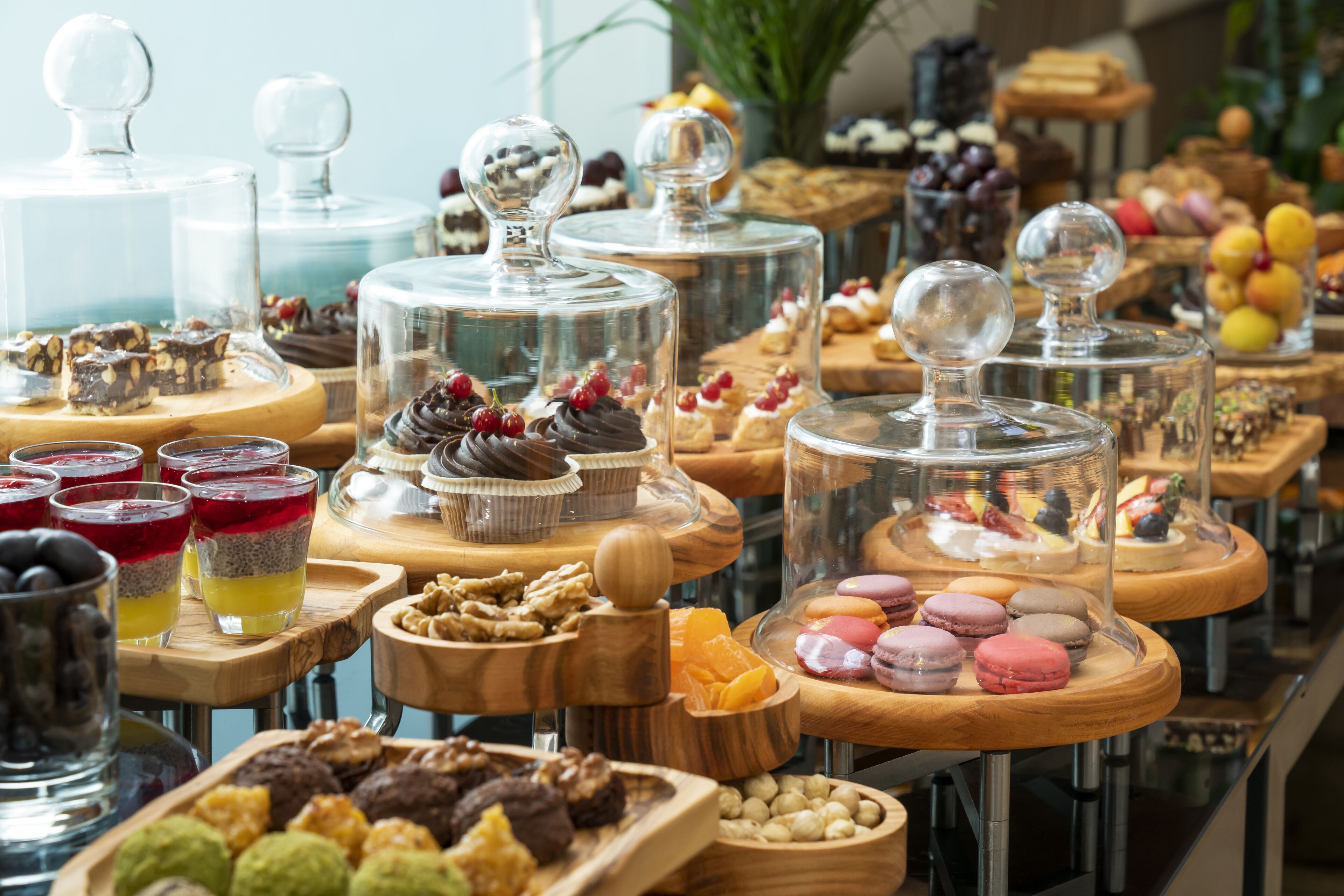 Un bufé con diversos postres y aperitivos, incluyendo macarons y opciones de chocolate, exhibidos en pedestales de madera con cubiertas de vidrio.