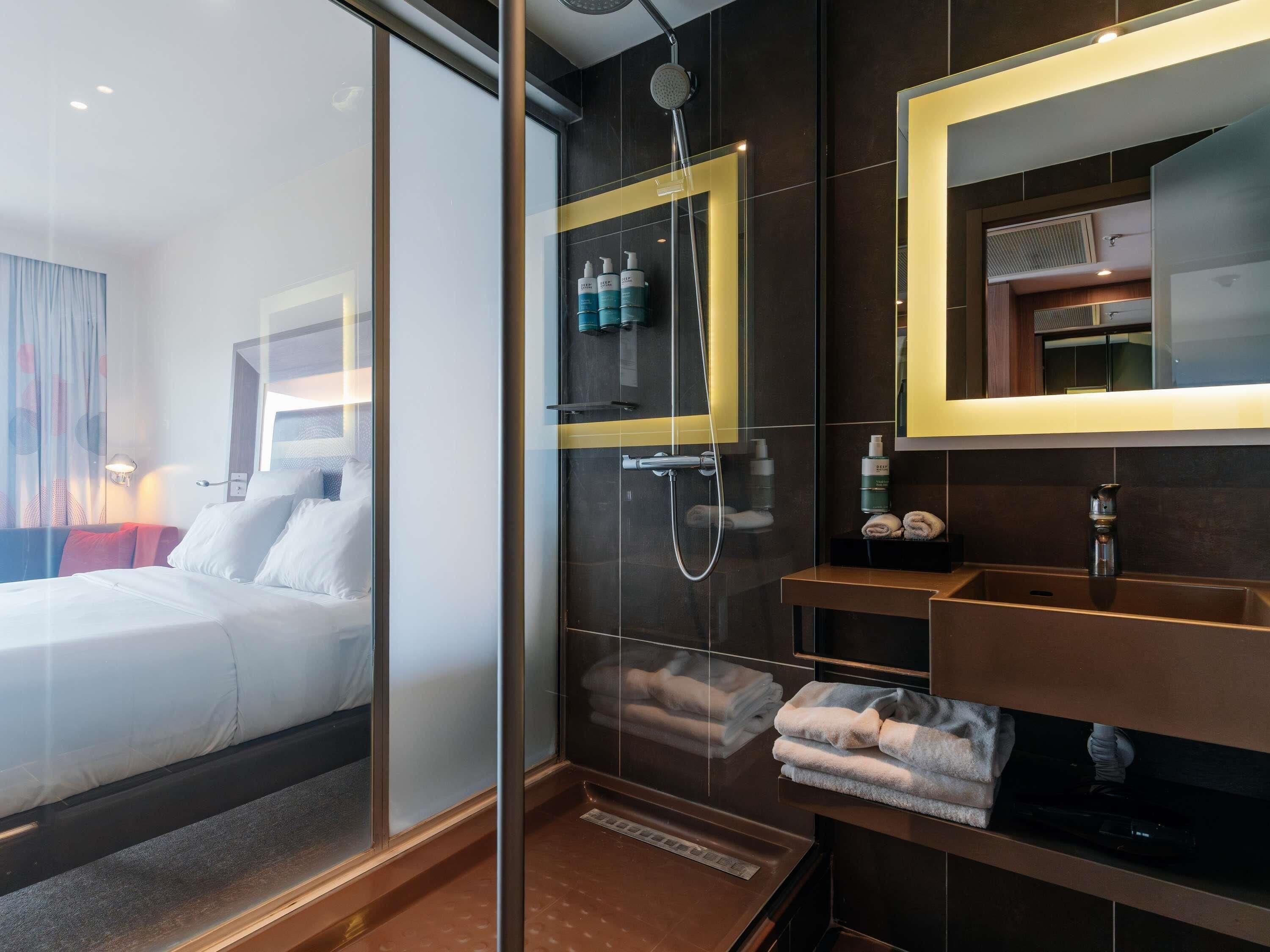 A modern hotel room with a glass partition, a large bed, a shower area, and a bathroom with a mirror and towel rack.